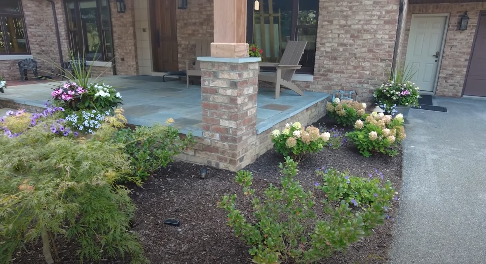 Elegant outdoor patio with stone fire pit, modern furniture, and professional landscaping featuring colorful flower beds and well-maintained lawn areas