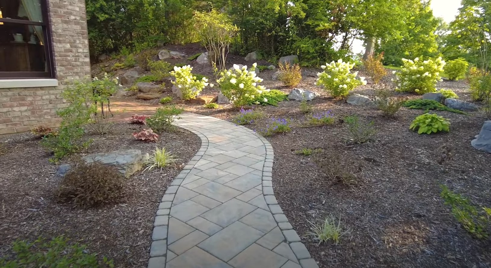 Curved stone walkway through beautifully landscaped garden with white hydrangeas, hostas, and natural stone accents